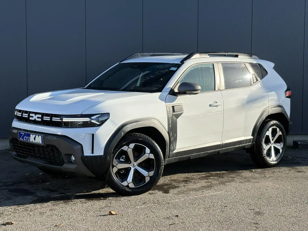 Vue 3/4 avant droit du Dacia Duster blanc glacier avec jantes alliage 18 pouces et barres de toit noires.