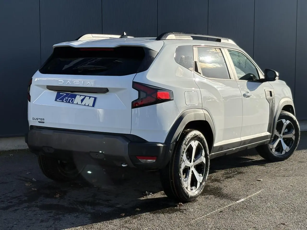 Vue 3/4 arrière droite du Dacia Duster blanc Glacier 2025 avec jantes alliage diamantées et barres de toit noires.