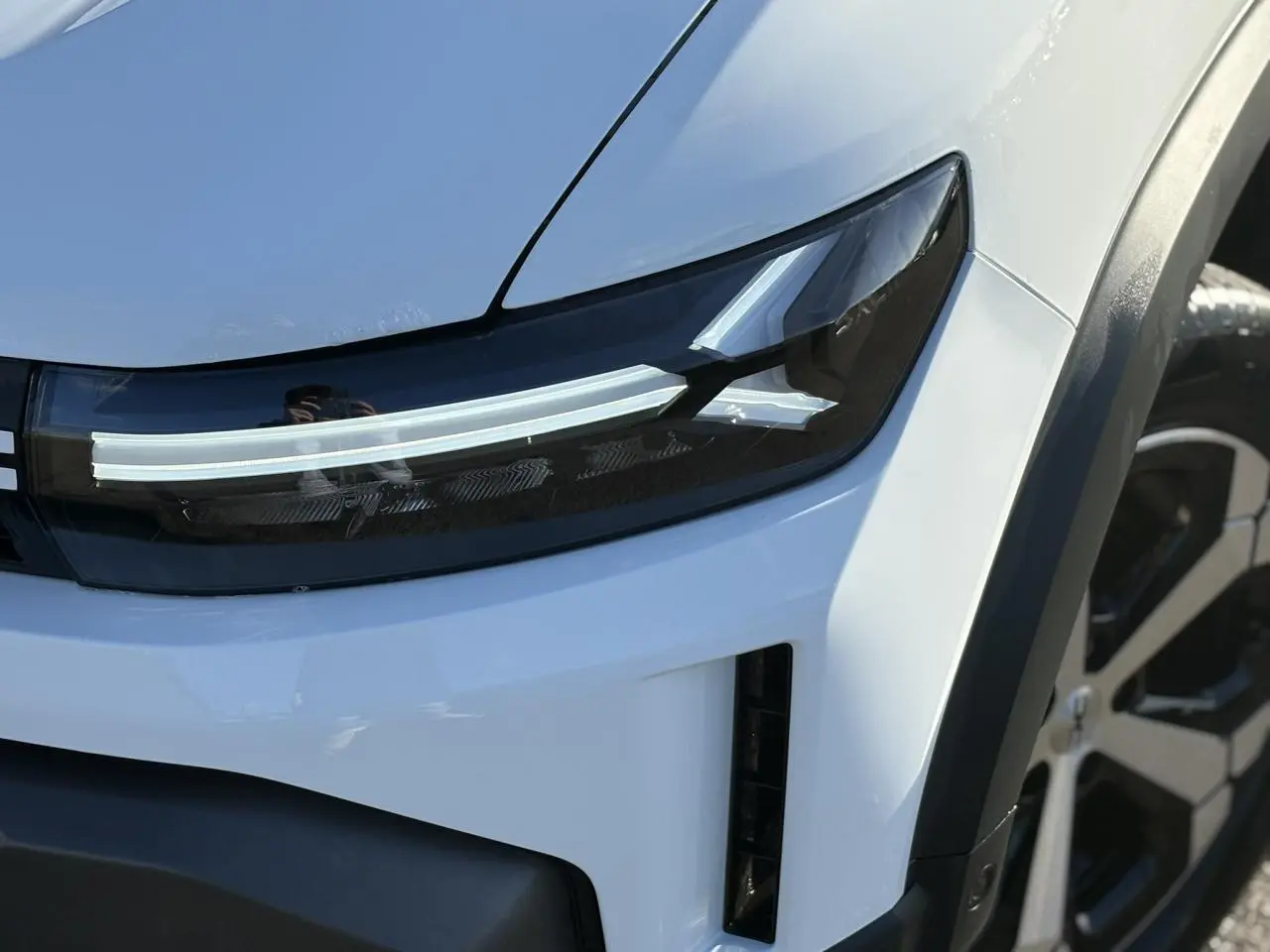 Gros plan sur le phare avant droit et la jante alliage diamantée d’un Dacia Duster blanc glacier 2025.