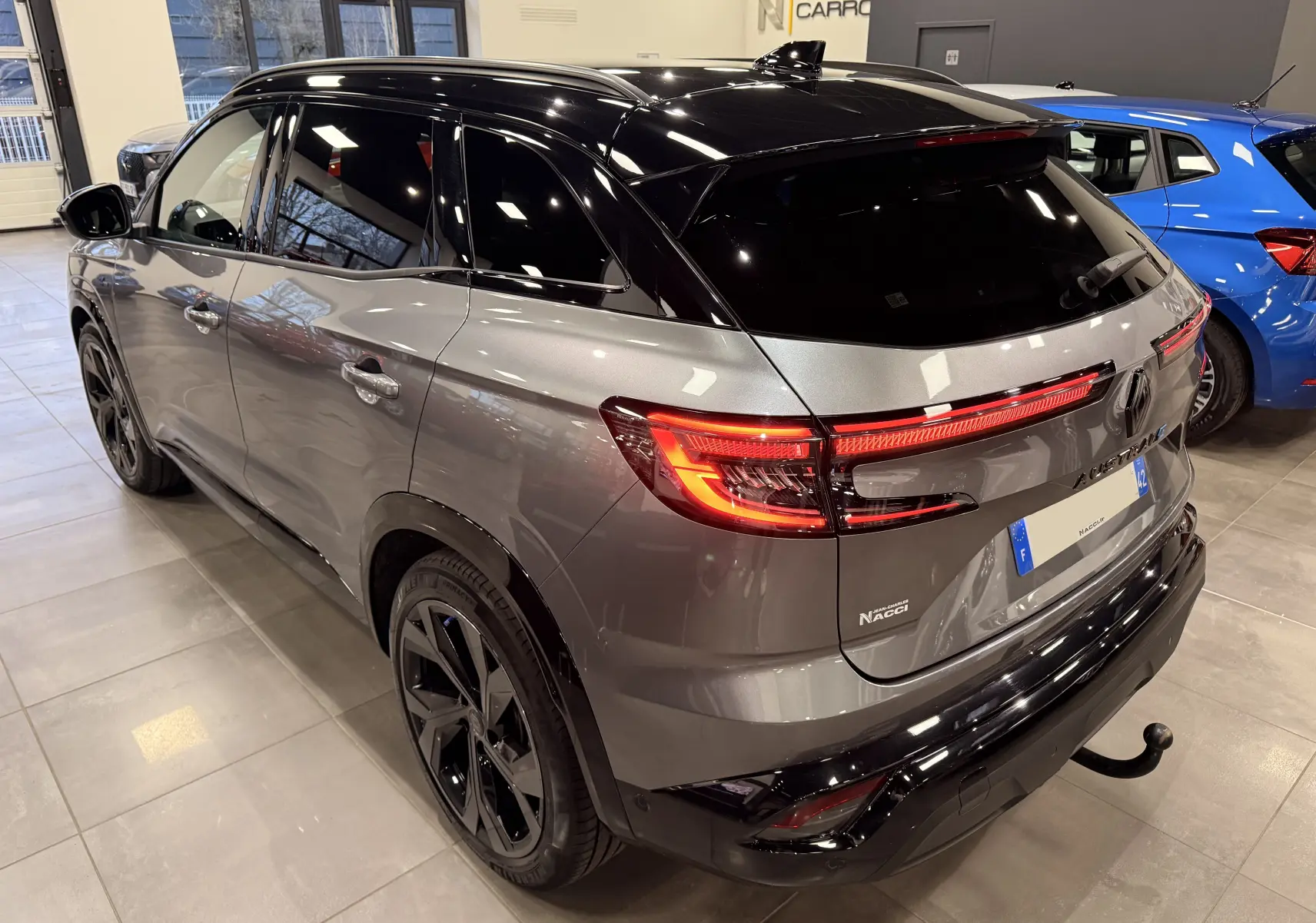 Vue 3/4 arrière droite du Renault Austral gris schist avec toit noir, feux LED allumés et attelage visible en intérieur showroom.