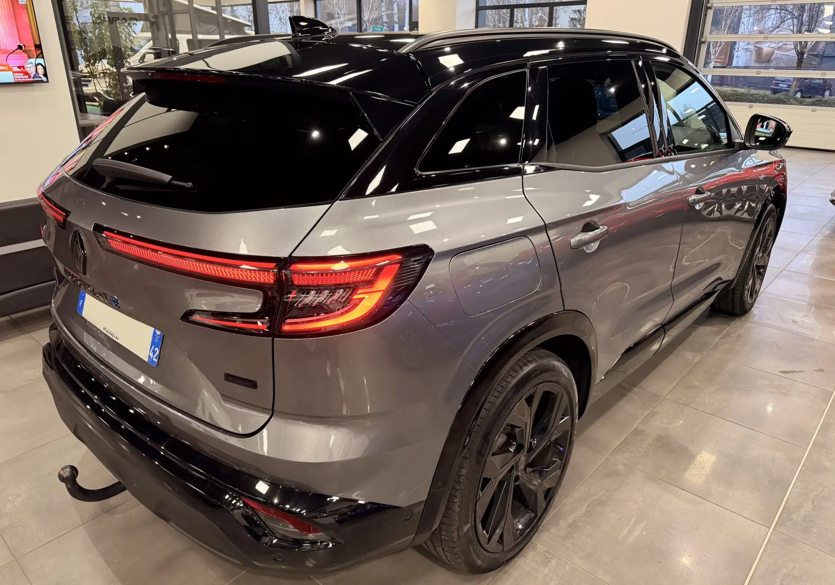 Vue 3/4 arrière droite du Renault Austral gris schist avec toit noir, feux arrière LED allumés en intérieur showroom.