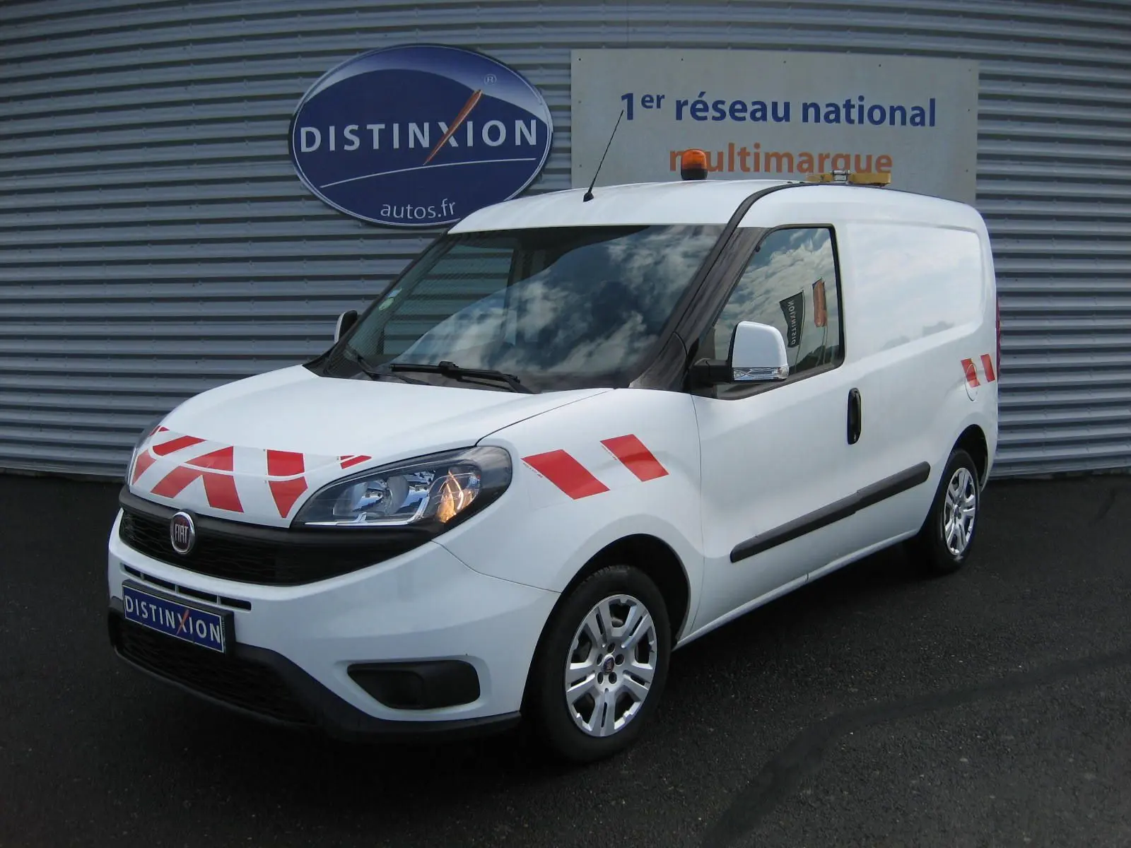 Vue 3/4 avant droit d’un FIAT Doblo Cargo blanc 2018 avec bandes rouges de signalisation et gyrophare orange sur le toit.