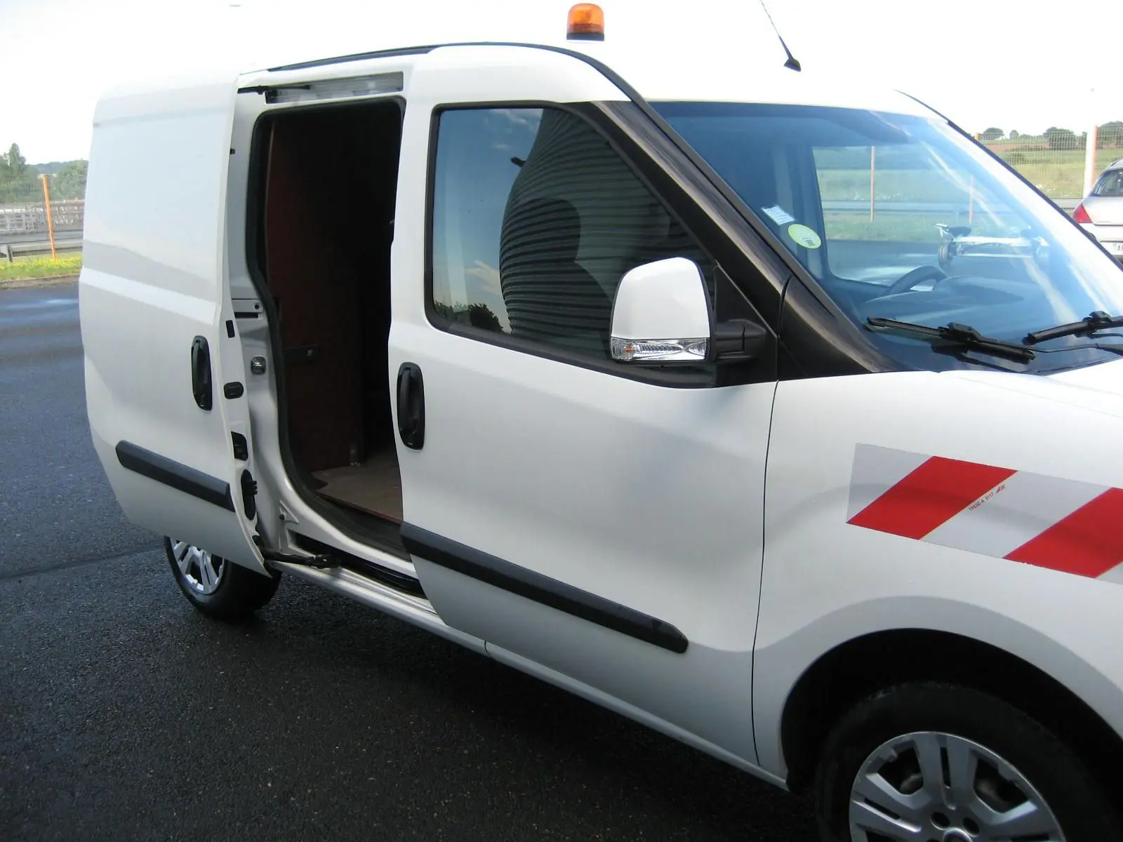 Vue de côté droit d'un FIAT Doblo Cargo blanc 2018 avec porte coulissante ouverte et bande réfléchissante rouge sur l'aile avant.