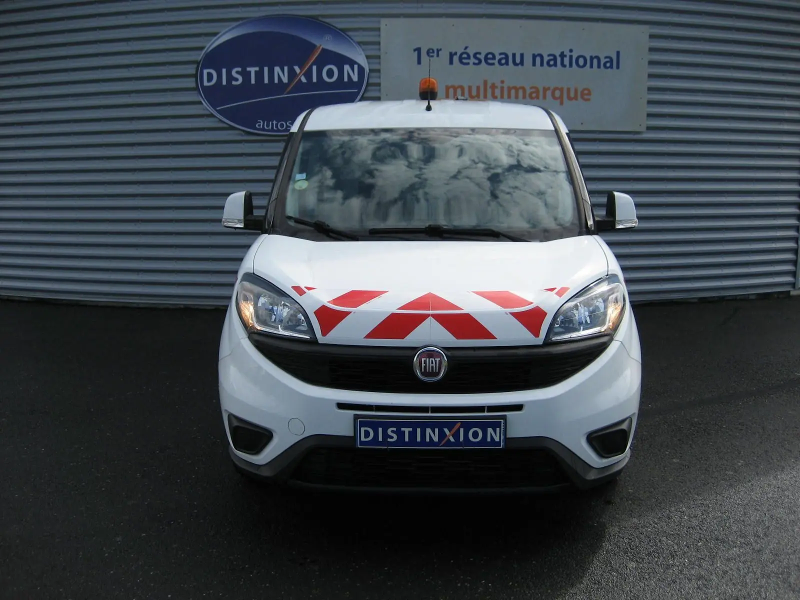 Vue frontale du FIAT Doblo Cargo blanc 2018 avec marquage rouge en chevrons sur le capot et gyrophare orange sur le toit.