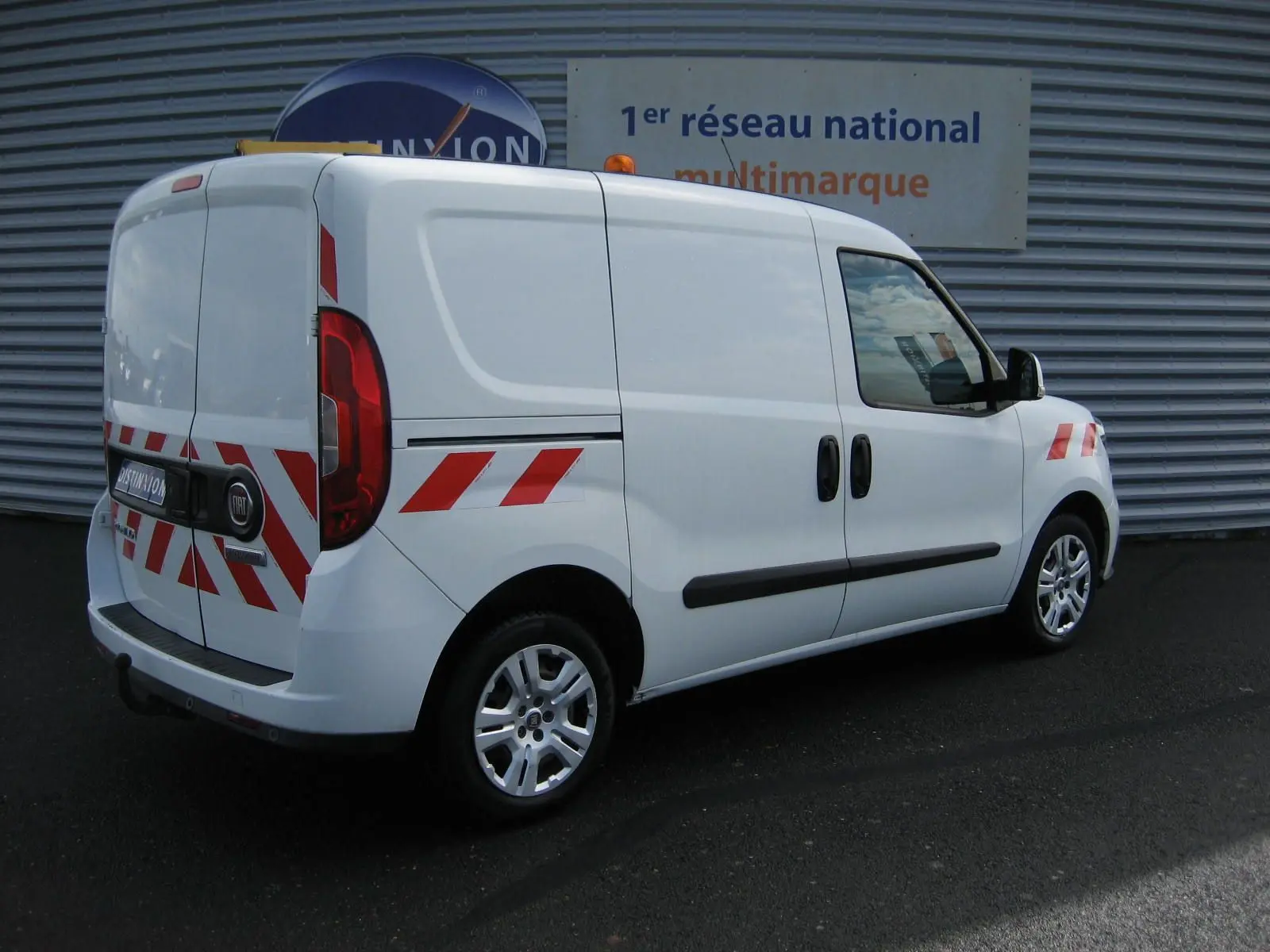 Fiat Doblo Cargo blanc vue 3/4 arrière droit avec bandes réfléchissantes rouges et blanches sur la carrosserie.