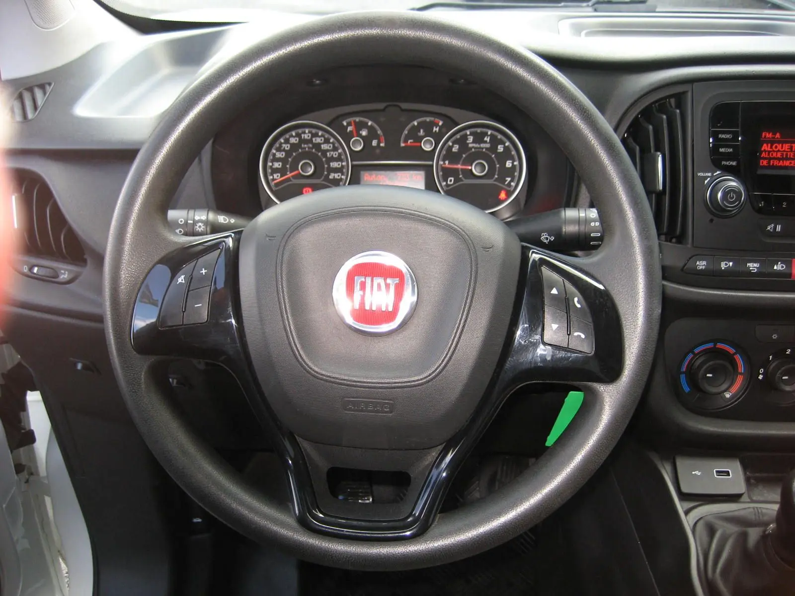 Vue rapprochée du volant noir avec logo FIAT rouge du Fiat Doblo Cargo blanc, tableau de bord et commandes visibles.