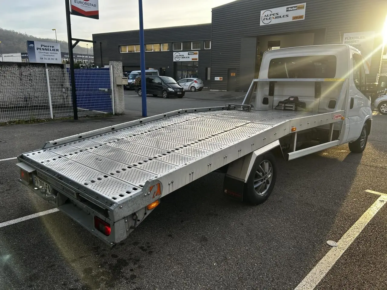 IVECO Daily blanc vu en 3/4 arrière droit, plateau métallique perforé pour transport, stationné en extérieur au soleil.