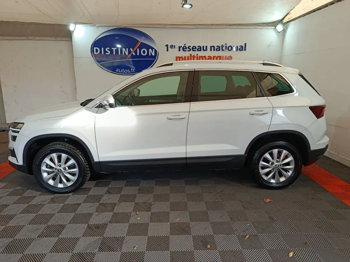 Vue latérale droite d'un Skoda Karoq blanc 2024 avec jantes alliage et barres de toit noires en intérieur showroom.