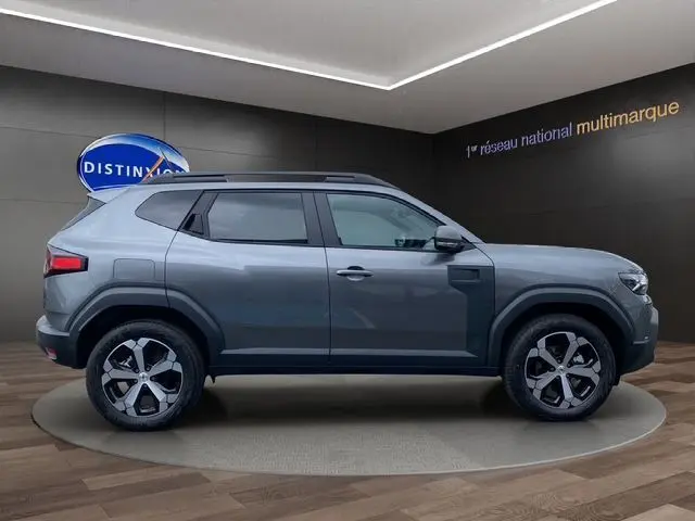 Profil côté droit du Dacia Duster 1.6 Hybrid 140 Journey gris schiste avec jantes alliage et barres de toit visibles.