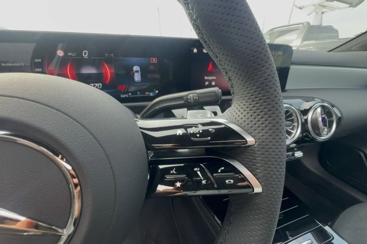 Gros plan sur le volant cuir perforé et les commandes multifonctions du tableau de bord de la Mercedes Classe A blanche.