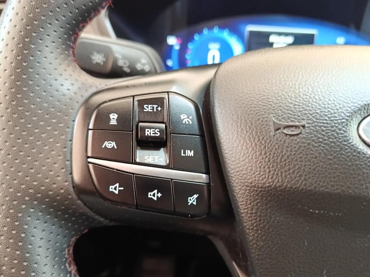Gros plan sur les commandes gauche du volant en cuir noir perforé du Ford Kuga hybride 2023, avec tableau de bord digital bleu en arrière-plan.