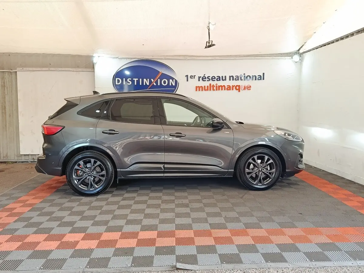 Ford Kuga hybride gris foncé vu de profil côté gauche, avec jantes noires et toit noir dans un showroom.