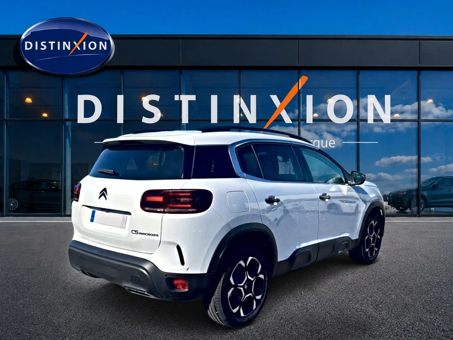 Vue 3/4 arrière droite d'un Citroën C5 Aircross blanc avec jantes alu et barres de toit devant un showroom.
