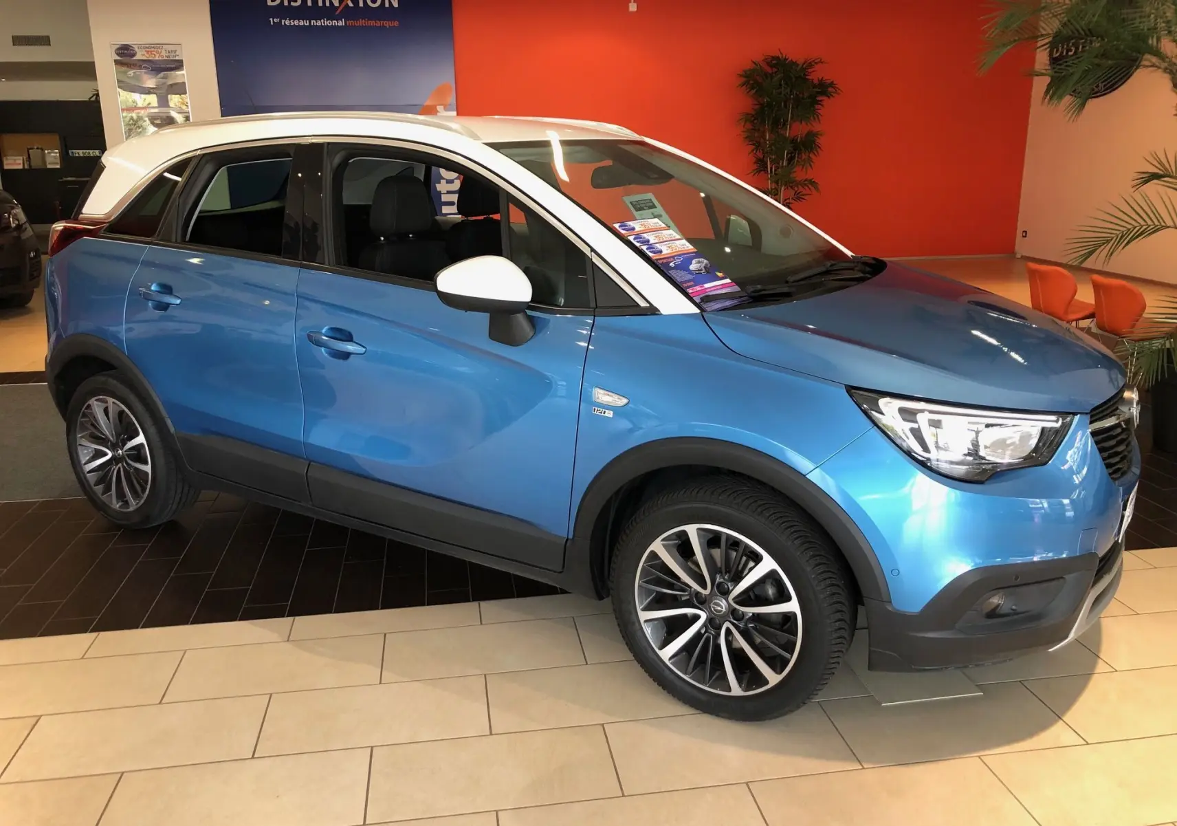 Opel Crossland X bleu métal en vue 3/4 avant droit, toit et rétroviseurs blancs, intérieur noir visible.