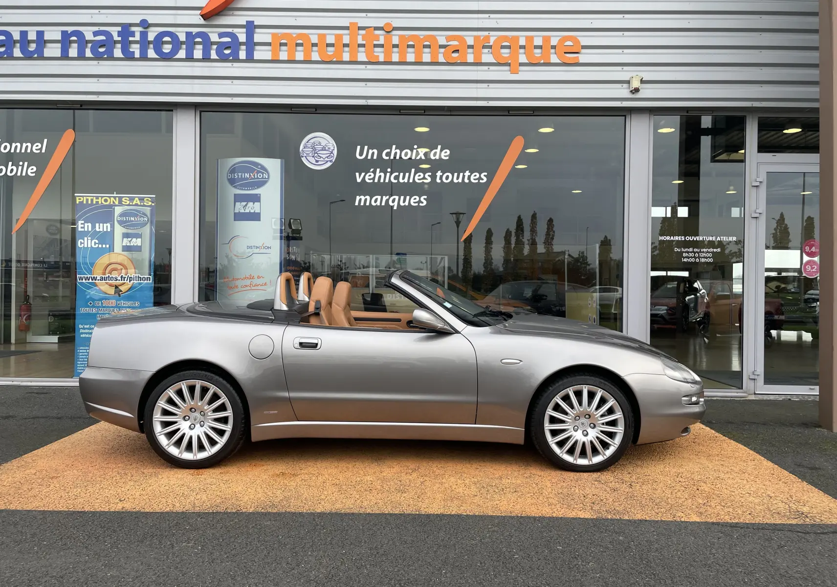 Profil droit d'une Maserati Spyder gris Nuvolari cabriolet de 2002 avec intérieur cuir beige, stationnée devant un showroom multimarque.