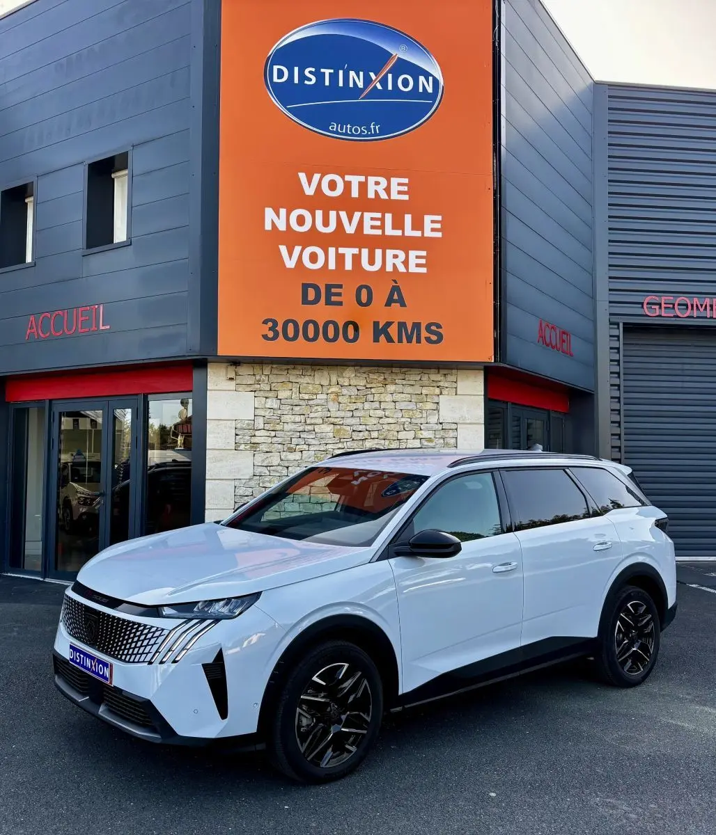 Peugeot 5008 blanc vue 3/4 avant droit, avec jantes noires et calandre distinctive, devant un bâtiment commercial.