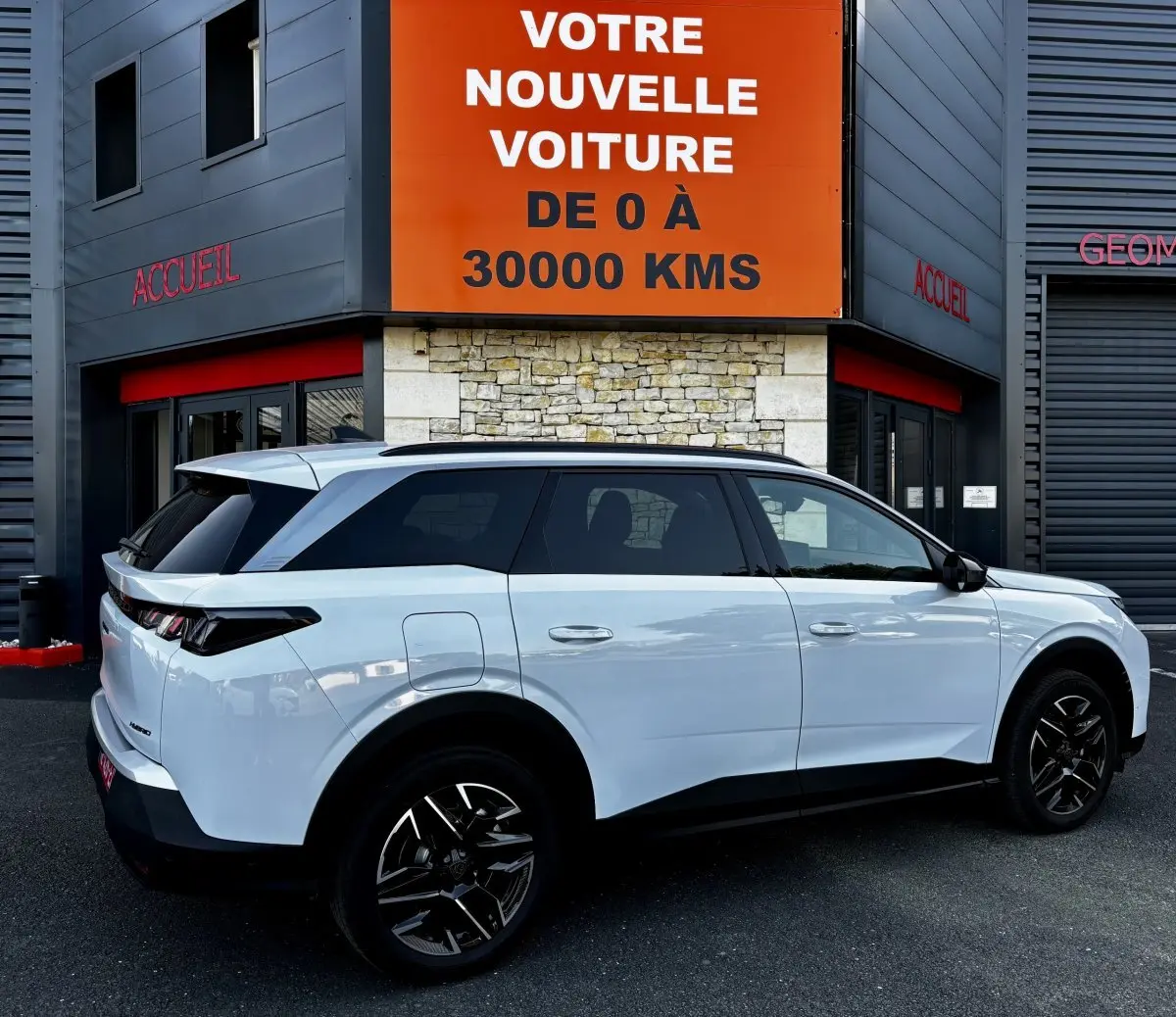 Peugeot 5008 hybride blanc vue de profil droit, stationnée devant un bâtiment avec panneau orange "Votre nouvelle voiture".