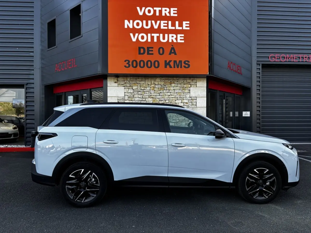Profil droit du Peugeot 5008 blanc hybride 2025, avec jantes noires et vitres teintées, devant un bâtiment commercial.