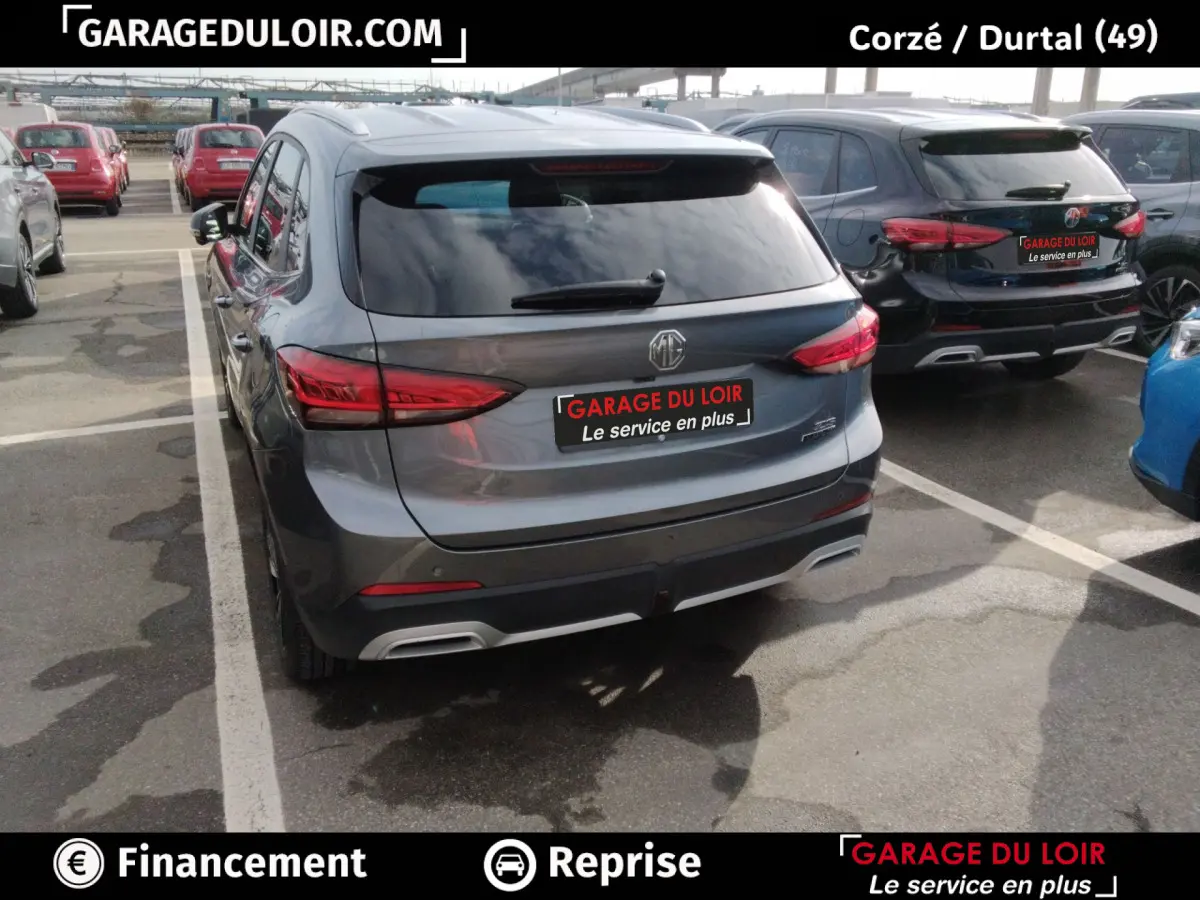Vue 3/4 arrière d’un MG ZS gris clair hybride, avec feux arrière LED et logo MG visible sur un parking.