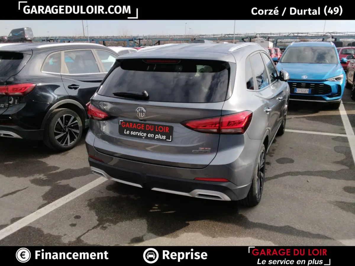 Vue 3/4 arrière d'un MG ZS gris clair hybride, avec feux arrière LED et logo MG visible sur un parking.