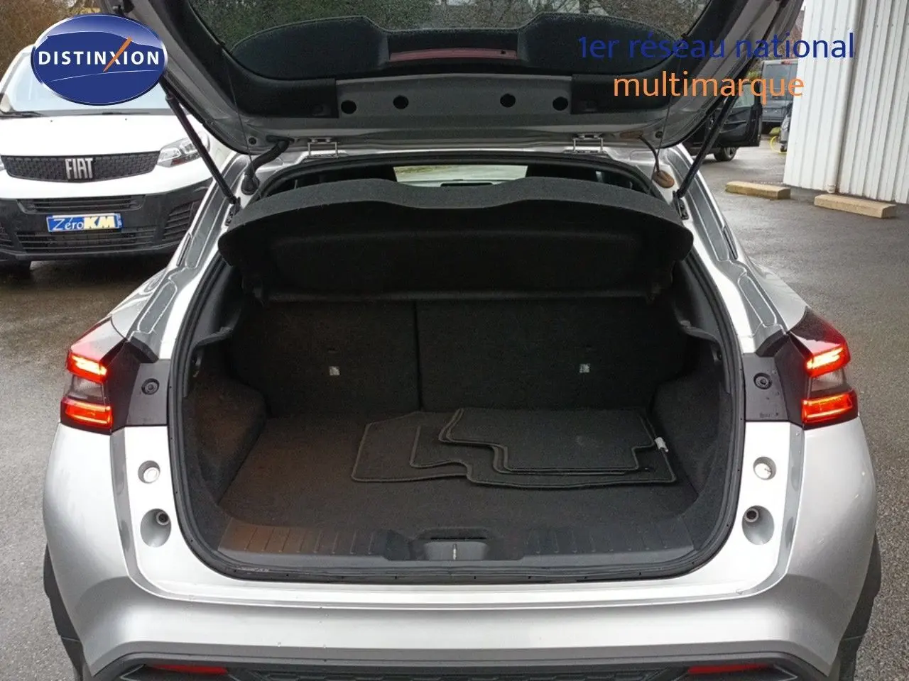 Vue arrière d'un Nissan Juke gris platine 2020 avec coffre ouvert et tapis de sol visibles, feux arrière allumés.