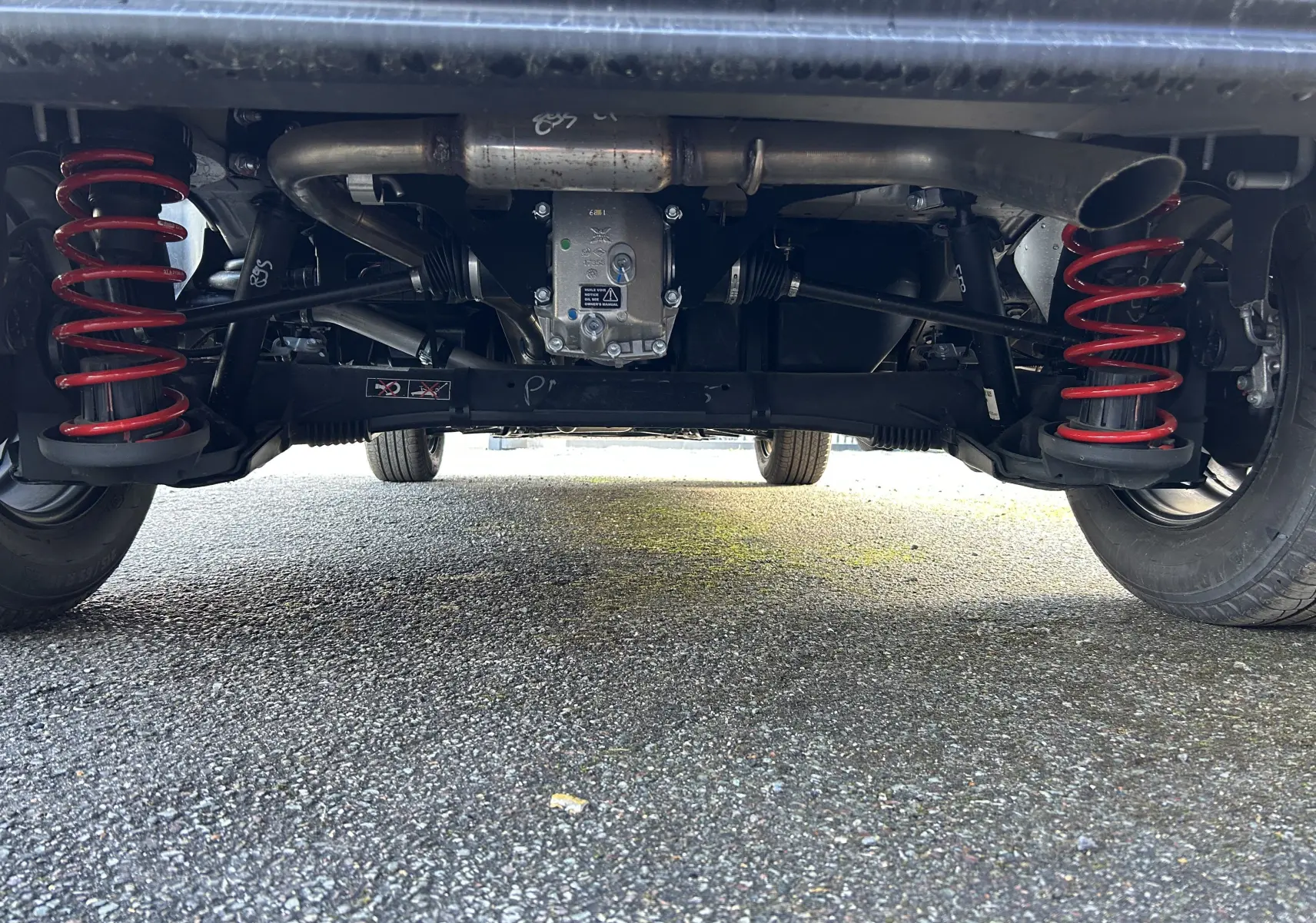 Vue en contre-plongée sous le train arrière du DANGEL Peugeot Partner gris avec suspension renforcée et échappement visible.