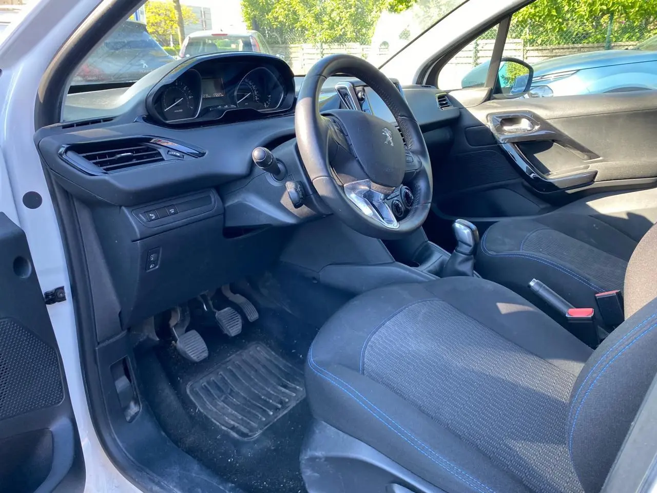 Vue intérieure côté conducteur de la Peugeot 208 blanche 2019, volant compact et sièges tissu avec surpiqûres bleues.