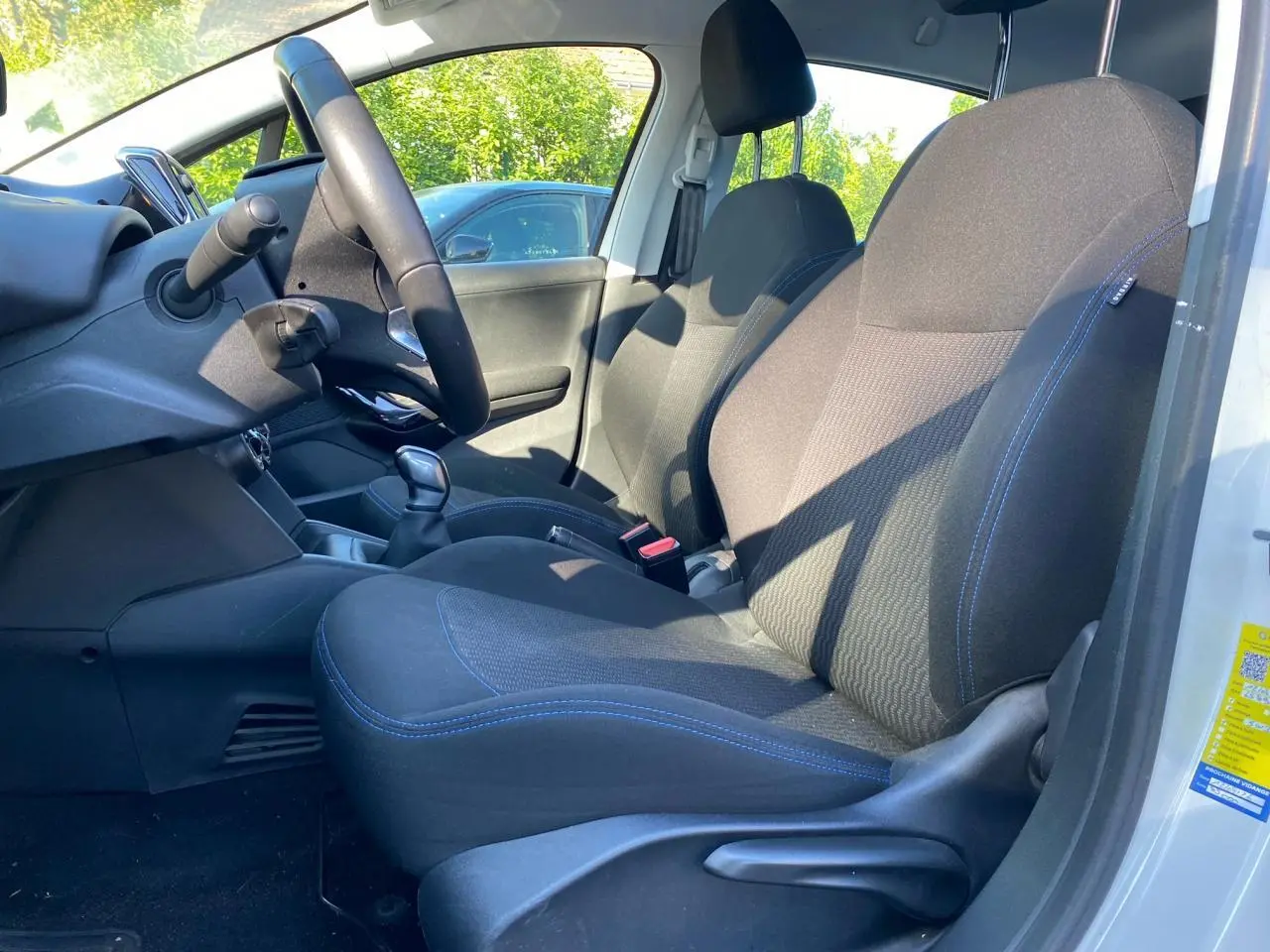 Vue intérieure côté conducteur de la Peugeot 208 blanche, montrant les sièges noirs avec surpiqûres bleues et le volant compact.