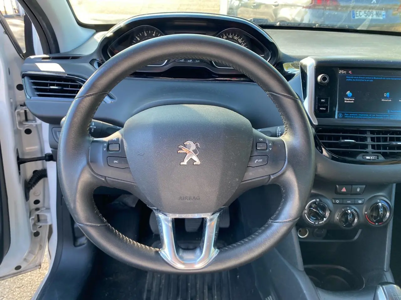 Vue intérieure centrée sur le volant Peugeot noir du 208 blanc, avec tableau de bord et écran tactile visible à droite.