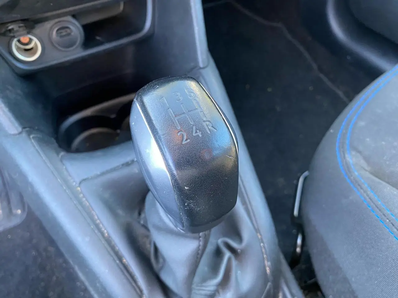 Gros plan sur le levier de vitesses manuel noir de la Peugeot 208 blanche, avec siège tissu gris et surpiqûres bleues visible à droite.