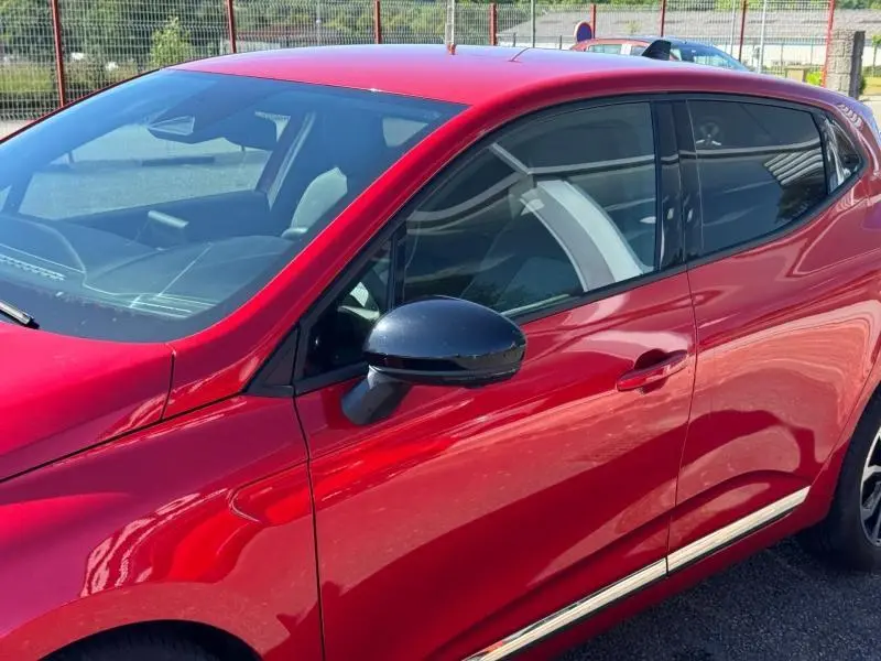 Vue de côté droit avant d'une Renault Clio rouge 2025 avec rétroviseur noir et vitres teintées.