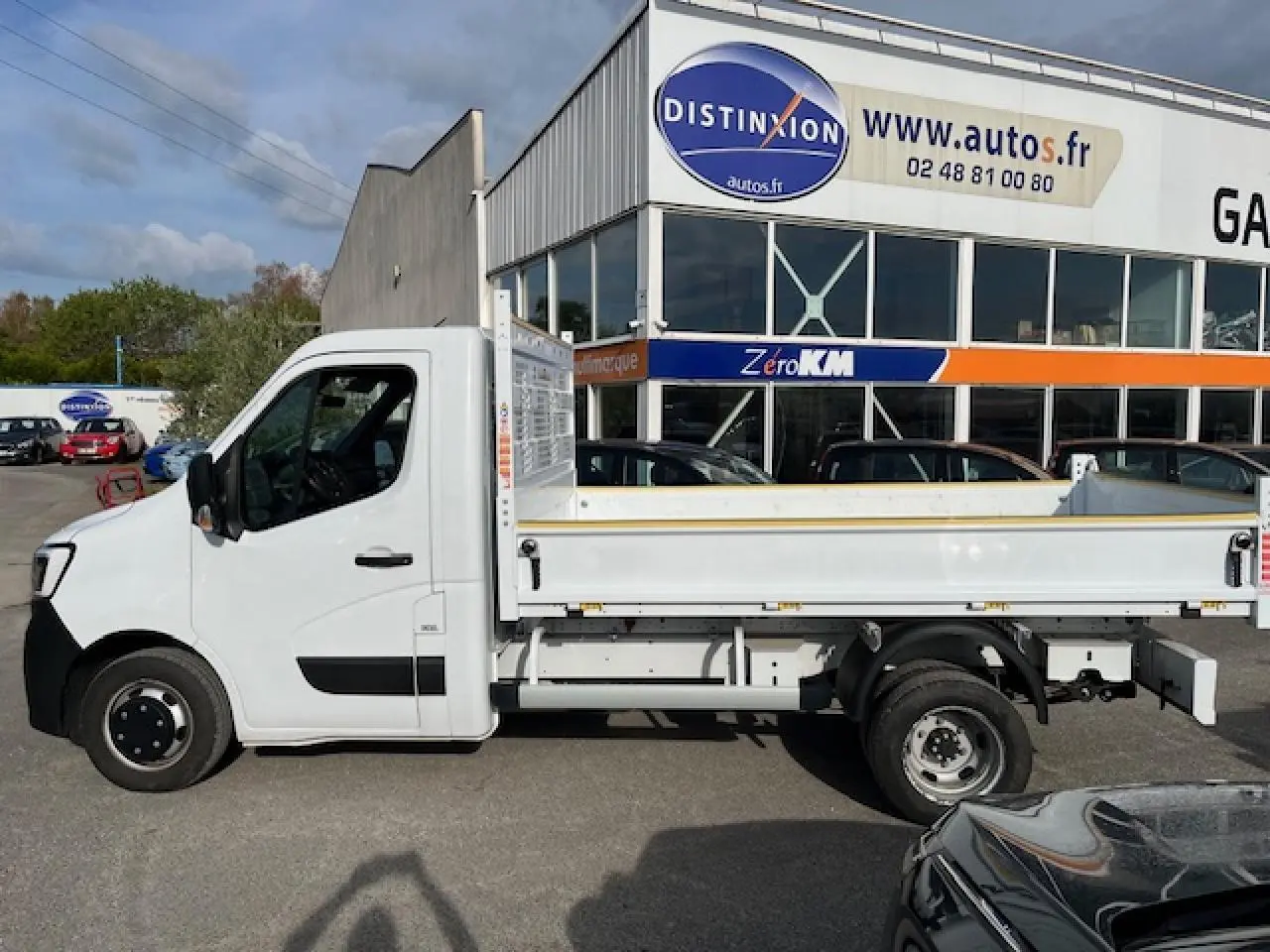 Vue latérale gauche d’un Renault Master blanc 2024 avec benne basculante, stationné devant un bâtiment commercial.