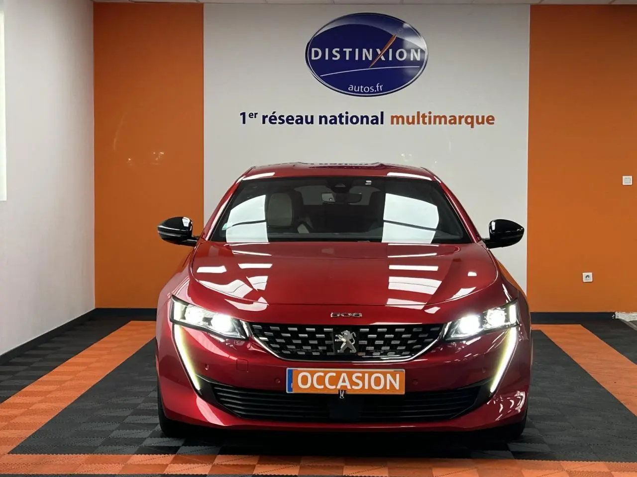 Vue frontale d'une Peugeot 508 Hybrid rouge clair avec phares allumés et plaque occasion dans un showroom.