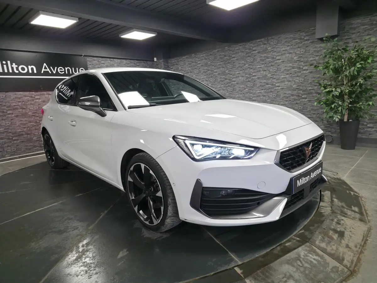 Vue 3/4 avant droite d'une CUPRA Leon blanche Nevada avec jantes noires et feux LED allumés en showroom.