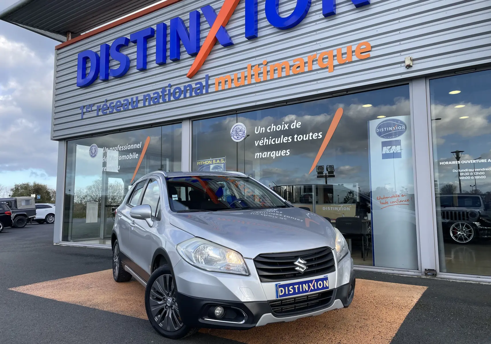 Suzuki SX4 S-Cross gris métallisé, vue 3/4 avant droit, avec projecteurs allumés et calandre noire distinctive.