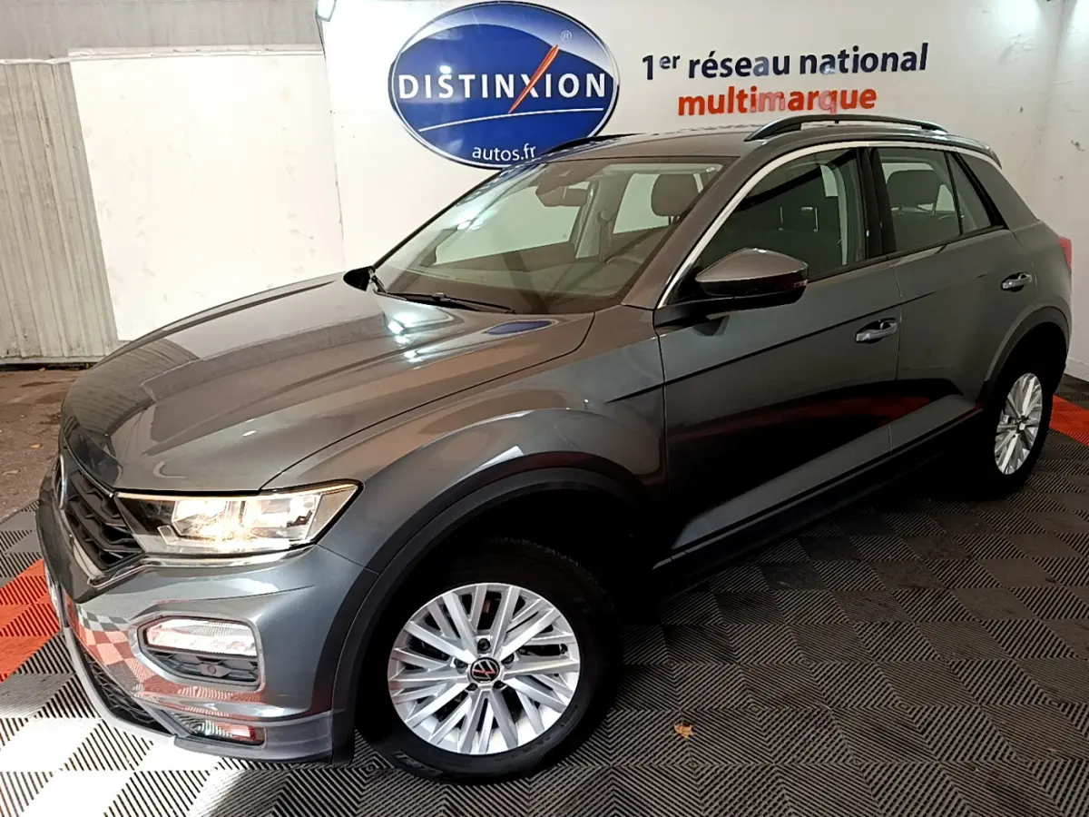 Volkswagen T-Roc gris foncé vue 3/4 avant droit, avec jantes argentées et phares allumés en intérieur showroom.