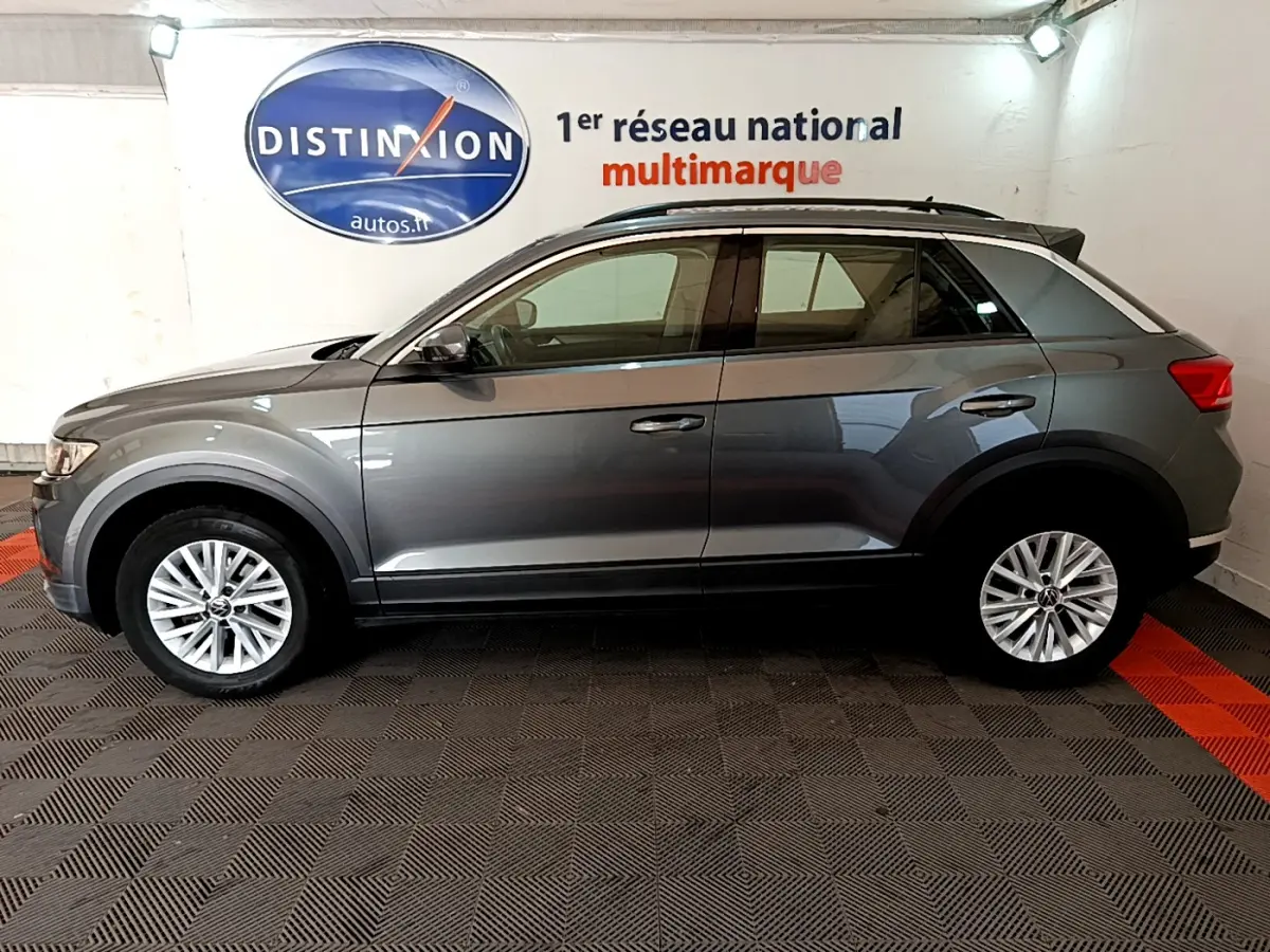 Profil côté gauche d'un Volkswagen T-Roc gris foncé 2021 dans un showroom avec logo Distinxion en arrière-plan.