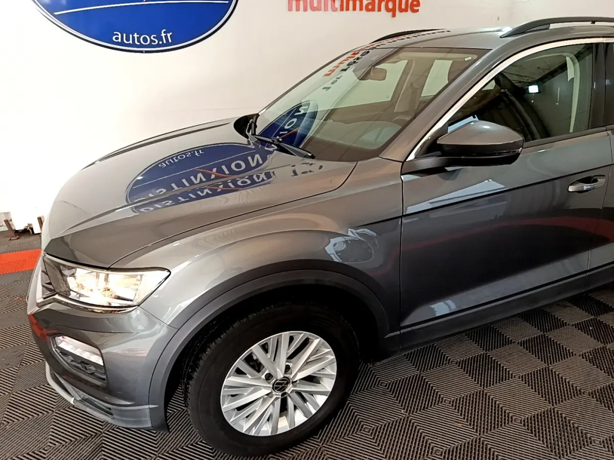 Vue 3/4 avant droit d'un Volkswagen T-Roc gris foncé avec jantes alliage et feux allumés dans un showroom.