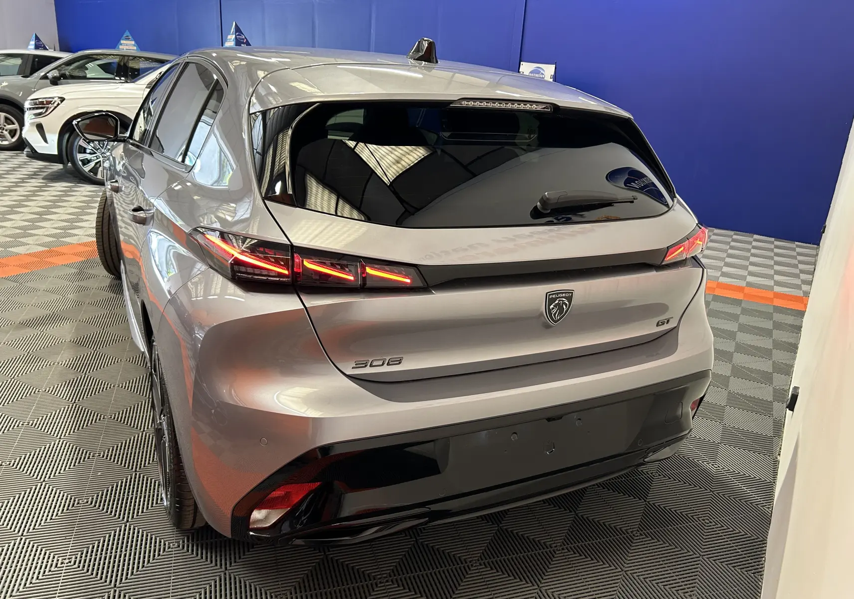 Vue 3/4 arrière droite de la Peugeot 308 GT 2026 gris Artense avec feux arrière LED allumés en intérieur showroom.