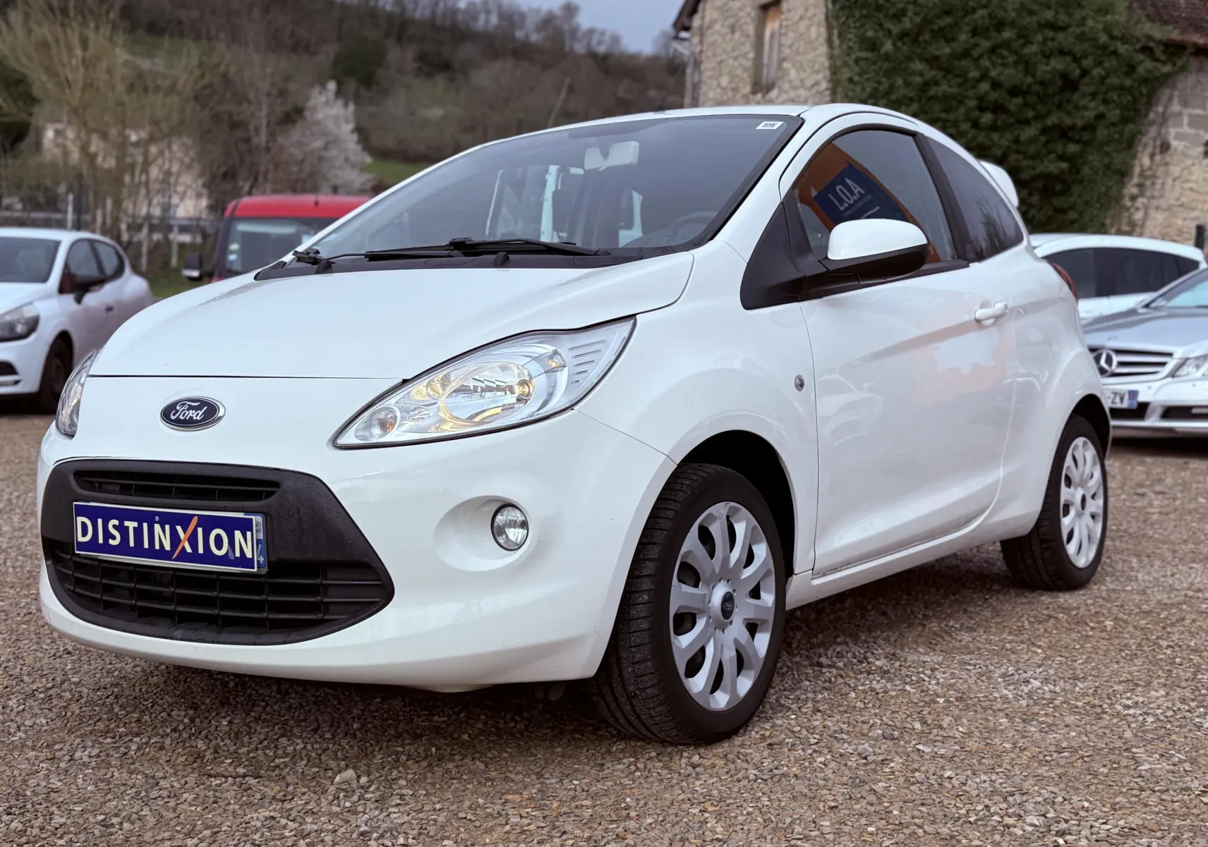 Ford Ka blanc vue 3/4 avant droit sur parking, avec phares allumés et jantes tôles blanches.
