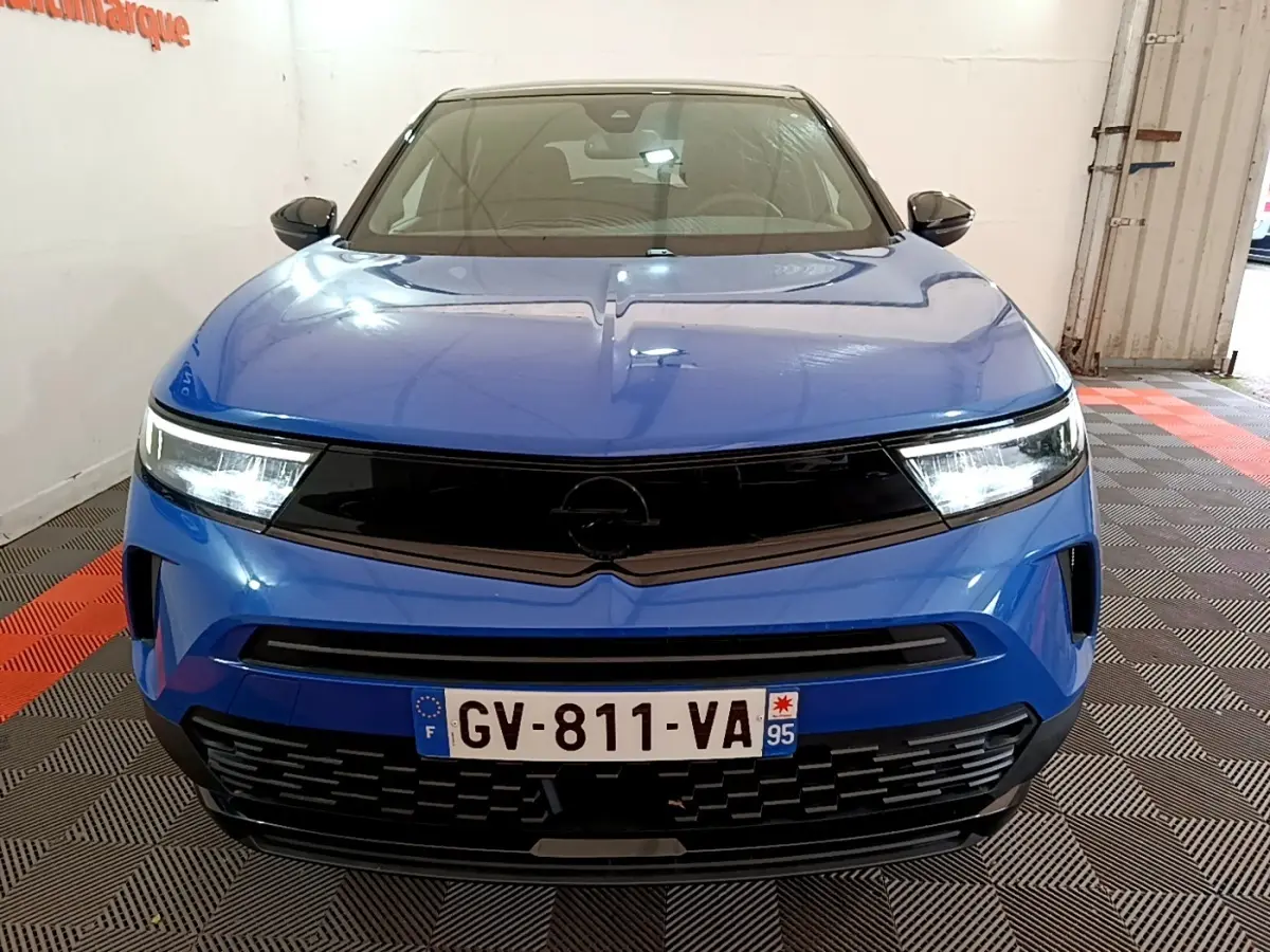 Vue avant d'un Opel Mokka bleu 2024 avec calandre noire et phares LED allumés dans un garage.