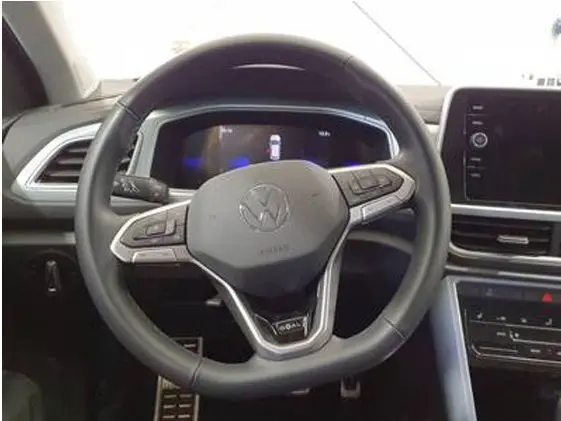 Vue rapprochée du volant noir du Volkswagen T-Roc 2025 avec commandes intégrées et tableau de bord numérique en arrière-plan.