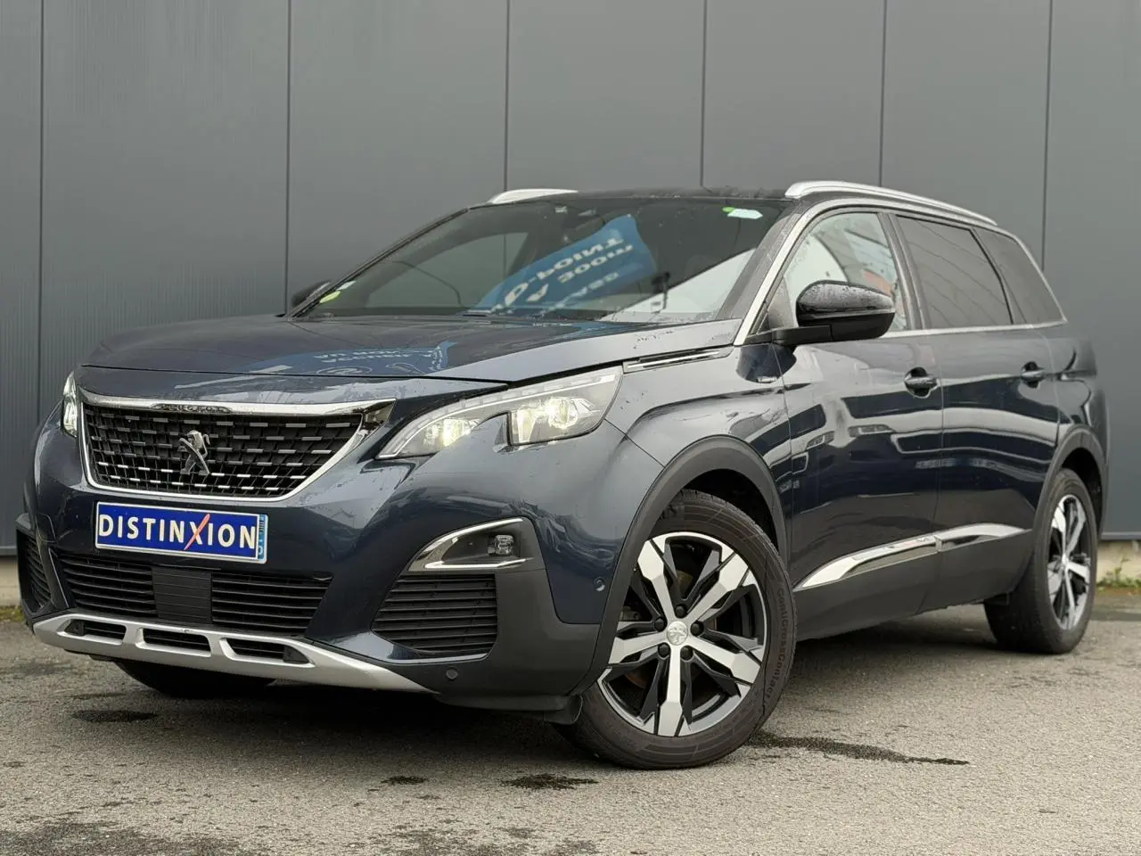 Vue 3/4 avant droit d’un Peugeot 5008 bleu foncé avec jantes alliage bi-ton et barres de toit argentées.