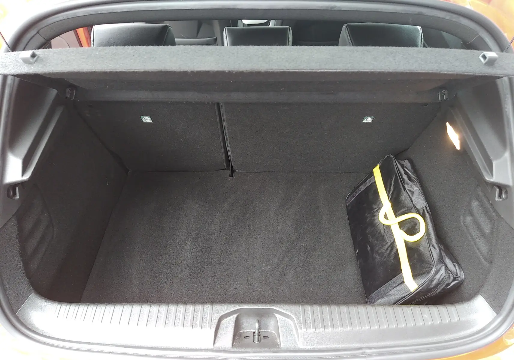 Coffre ouvert vu de l'arrière d'une Renault Clio V orange, avec un sac noir à bande jaune à l'intérieur.
