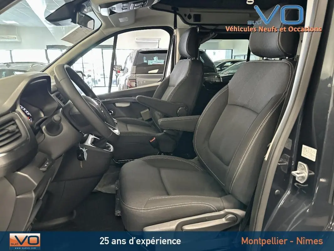 Intérieur côté conducteur du Renault Trafic Fourgon gris 2025, sièges tissu noirs avec accoudoirs et tableau de bord visible.