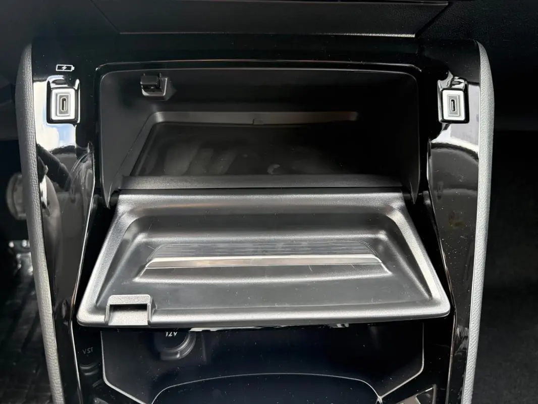Vue rapprochée du rangement central ouvert de la Peugeot 208 Hybrid 145 gris, avec prise 12V visible en dessous.