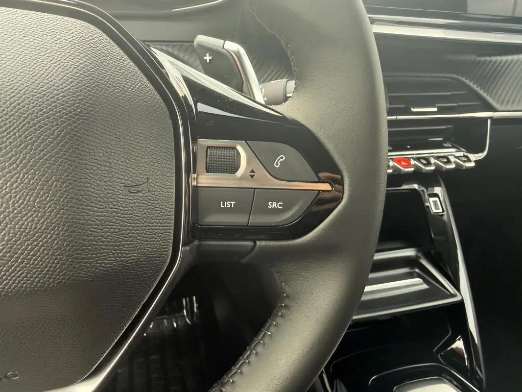 Gros plan sur le volant noir de la Peugeot 208 Hybrid 145 e-DCS6 Allure 2025 avec commandes intégrées et tableau de bord brillant.