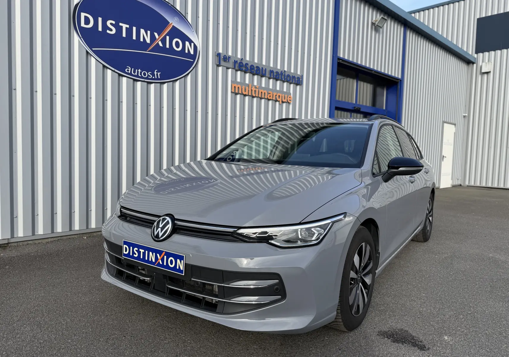 Volkswagen Golf SW 2.0 TDI gris lunaire vue 3/4 avant droit, devant un bâtiment avec enseigne Distinxion.