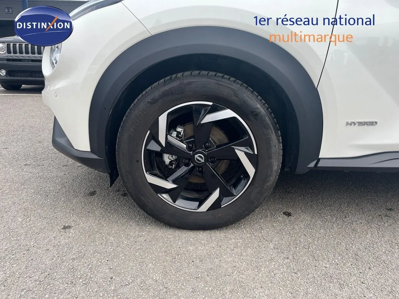 Gros plan sur la roue avant gauche noire et argentée du Nissan Juke blanc nacré hybride, avec badge Hybrid visible sur la carrosserie.