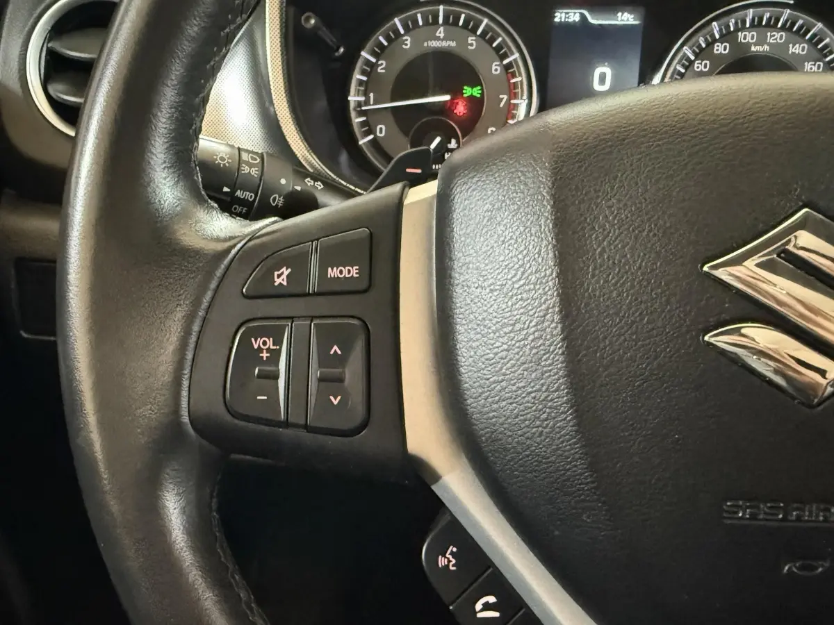 Gros plan sur le volant cuir noir du Suzuki Vitara 2022 avec commandes audio et tableau de bord éclairé en arrière-plan.